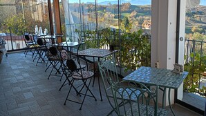 Ein überdachter Außenbereich mit Tischen und Stühlen, große Fenster mit Blick auf eine hügelige Landschaft.