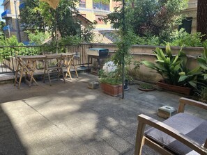 A rooftop patio with a wooden table and chairs, potted plants, and a view of buildings in the background.