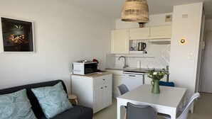 A compact kitchenette with a microwave, coffee maker, and sink. There is a dining table with chairs and a small sofa with patterned cushions. - F2 Apartment near beach with parking and pets allowed (Fort-Mahon-Plage)