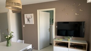 A modern living room with a flat-screen TV, a dining table with chairs, a decorative wall piece, and a door leading to another room. - F2 Apartment near beach with parking and pets allowed (Fort-Mahon-Plage)