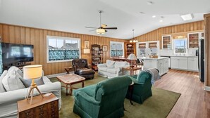 A spacious living room with a television, sofa, and armchairs, a dining area with a table and chairs, and a kitchen with white cabinets and a wooden ceiling. - Fish On - Channel Home Steps To The Beach (North Myrtle Beach)