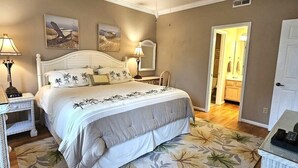 Une chambre à coucher comprenant un lit, des tables de chevet, un ventilateur de plafond et une porte donnant sur une salle de bain.