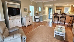 Un salon spacieux avec un canapé en cuir, une table basse et un téléviseur. Il y a un coin cuisine avec des placards en bois et un coin repas avec deux chaises.