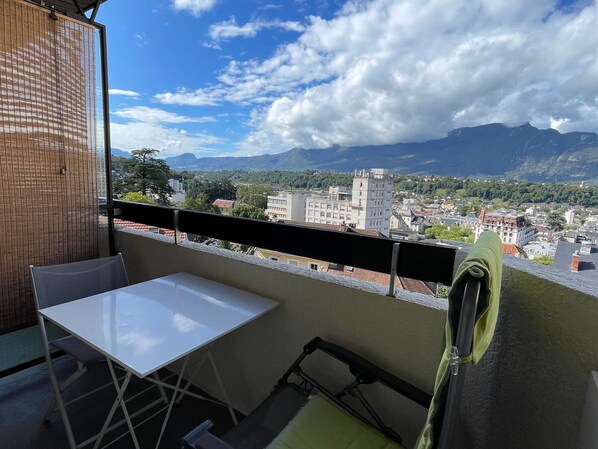 A balcony with a white table and chairs, overlooking a cityscape with mountains in the background.
