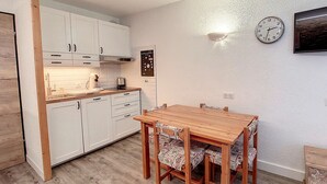 Une cuisine compacte avec des meubles blancs, une table à manger en bois et des chaises à motifs, ainsi qu’une horloge murale.