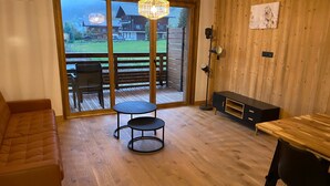 Un cuarto acogedor con piso de madera, un sofá de cuero marrón, una mesita redonda pequeña, un mueble de madera y un amplio ventanal corredero que da a una terraza exterior.