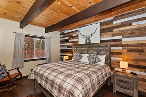 A bedroom with a wooden ceiling, a bed with plaid bedding, a nightstand with a lamp, and a window with curtains.