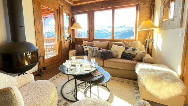 Ein gemütliches Zimmer mit Holzvertäfelung, einer Couch, einem runden Tisch mit Gläsern und einem großen Fenster mit Blick auf die Berge.