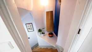 Una escalera estrecha con escalones de madera que conduce a un rellano. Un cuadro enmarcado cuelga de la pared, y hay una planta en maceta en el rellano.