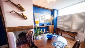 Un área de comedor con una mesa de madera, sillas y un jarrón azul. Hay una chimenea con un espejo encima y una ventana con persianas.