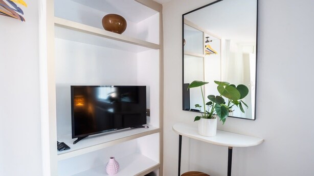 Una sala moderna con una televisión de pantalla plana sobre un estante blanco, una pequeña planta en maceta y un taburete redondo de madera.