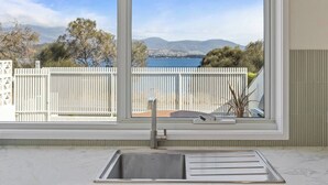 A kitchen with a large window overlooking a white fence and a scenic view of a lake and mountains.