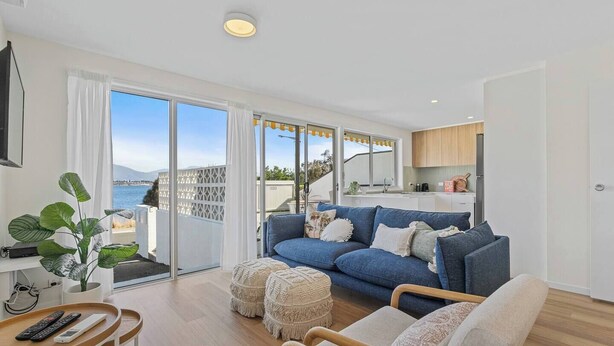 A modern living room with a blue sofa, wooden chairs, and a small table with remotes. A large window offers a view of the sea and mountains.