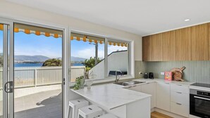 A modern kitchen with a view of the sea, a dining area with stools, and a wooden cabinet.