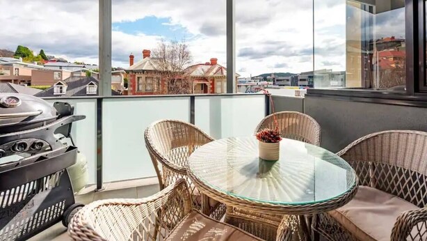 En balkong med flätade stolar och ett glasbord, med utsikt över ett bostadsområde med hus och en klar himmel.
