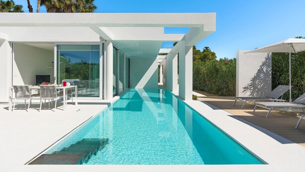 A modern poolside area with a clear pool, white outdoor furniture, and a glass-enclosed patio. - Beautiful modern villa with private pool, just steps from the sea in Fanusa, Syracuse. (Siracusa)