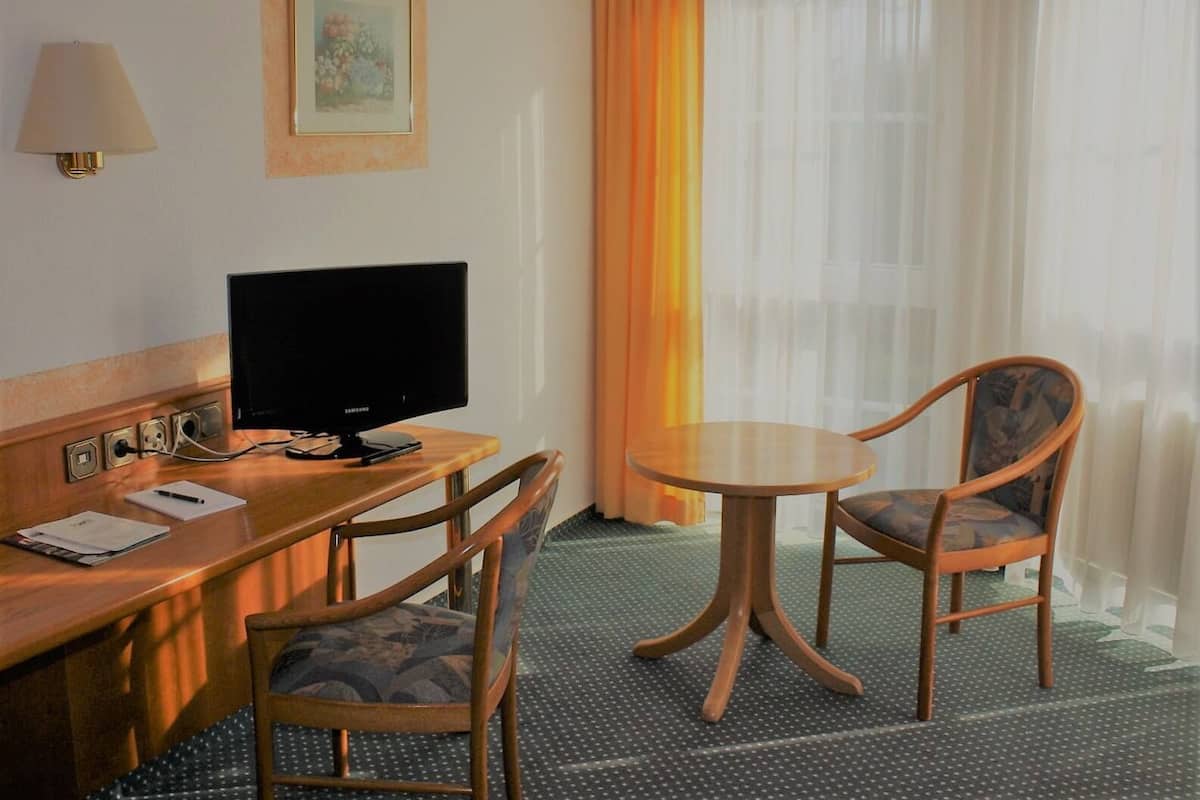 Ein Hotelzimmer mit einem Holztisch, einem Fernseher, zwei Sesseln, einem kleinen Tisch, einer Lampe und einem Fenster mit Vorhängen.
