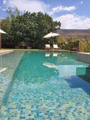 A clear blue pool with tiled flooring, surrounded by lounge chairs and umbrellas, with a view of a mountainous landscape in the background. - Le Figuier du lac (Bine el Ouidane)