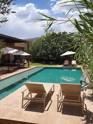 A swimming pool with two wicker lounge chairs in the foreground, surrounded by greenery and a covered seating area with umbrellas. - Le Figuier du lac (Bine el Ouidane)