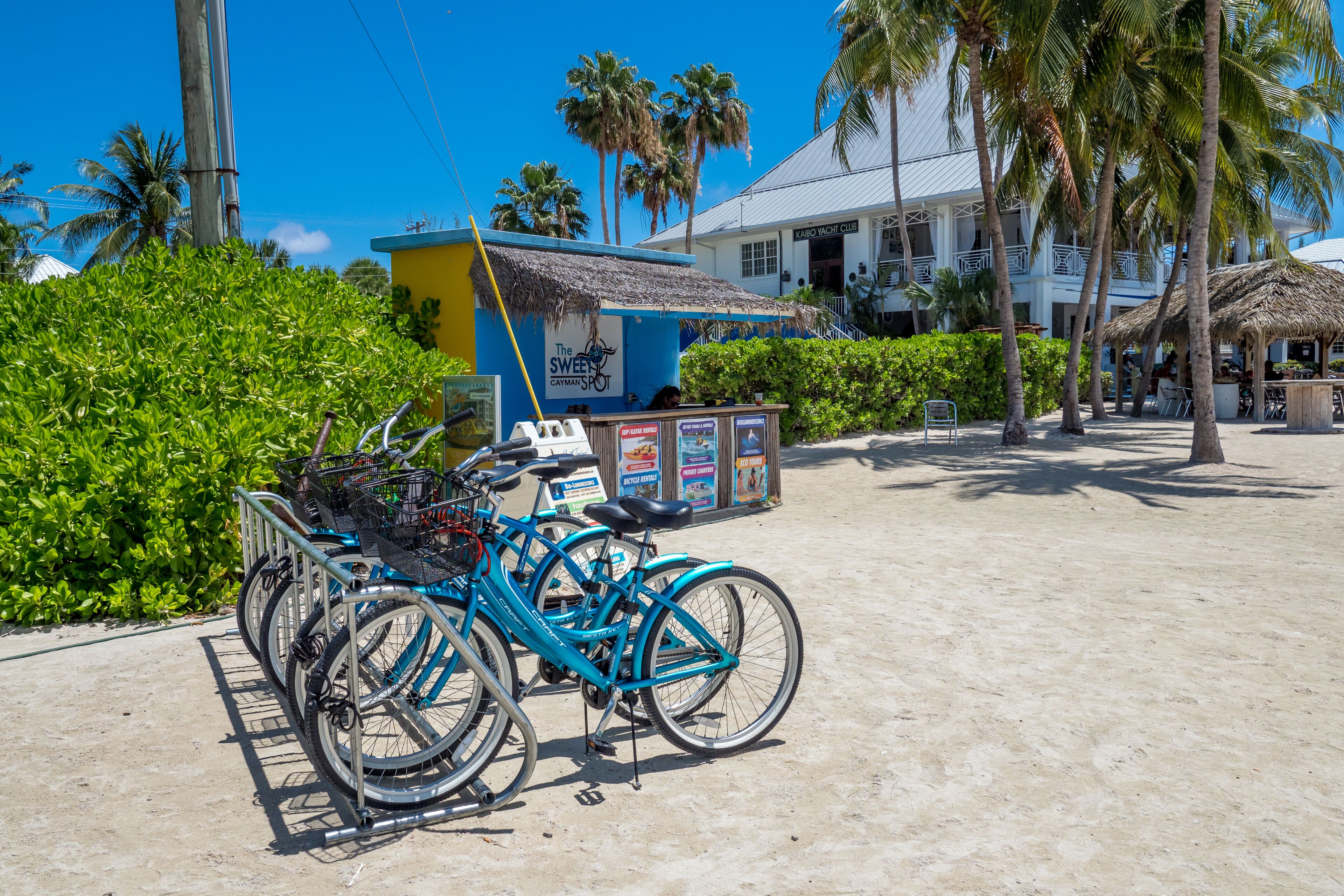 A bicycle rental station with a blue bicycle, a sign for 'The Sweet Spot Cayman', and a building with a thatched roof in the background.