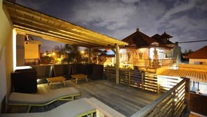 Una terraza de madera con sillones de descanso y un área de estar cubierta, con vista a un edificio tradicional de tejas.
