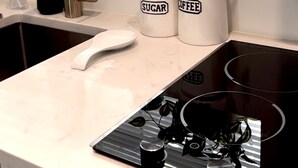 Une cuisine moderne avec un évier, un robinet et une cuisinière. Sur le comptoir, on trouve deux récipients blancs étiquetés « SUCRE » et « CAFÉ ». Une plante est posée sur le rebord de la fenêtre, et un rouleau d’essuie-tout est accroché au mur.