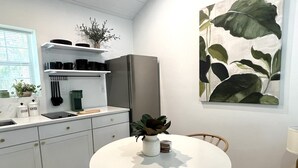 Une cuisine moderne avec des armoires blanches, un réfrigérateur en acier inoxydable et une table à manger avec des chaises. On peut voir une plante en pot sur le comptoir et un tableau représentant des feuilles sur le mur.