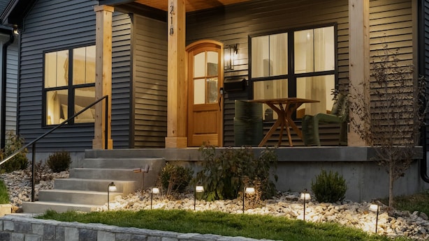 A modern house with a dark exterior, a front porch with a wooden railing, and a stone retaining wall.