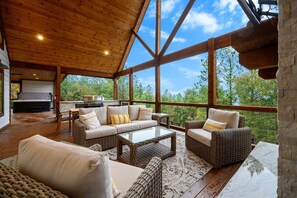 A spacious covered patio with wicker furniture, a glass coffee table, and a stone fireplace. - Magical 3-bed cabin with pool table & hot tub. Greet the day with wooded mountain views, relax by the firepit, then unwind in the bubbling spa. (Broken Bow)