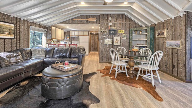 A spacious living area with a leather sofa, a round ottoman, and a dining table with white chairs. The room features a high ceiling with a fan, wooden panel walls, and a cowhide rug. - Serene and Secluded Walk Up in the Woods (Ligonier)