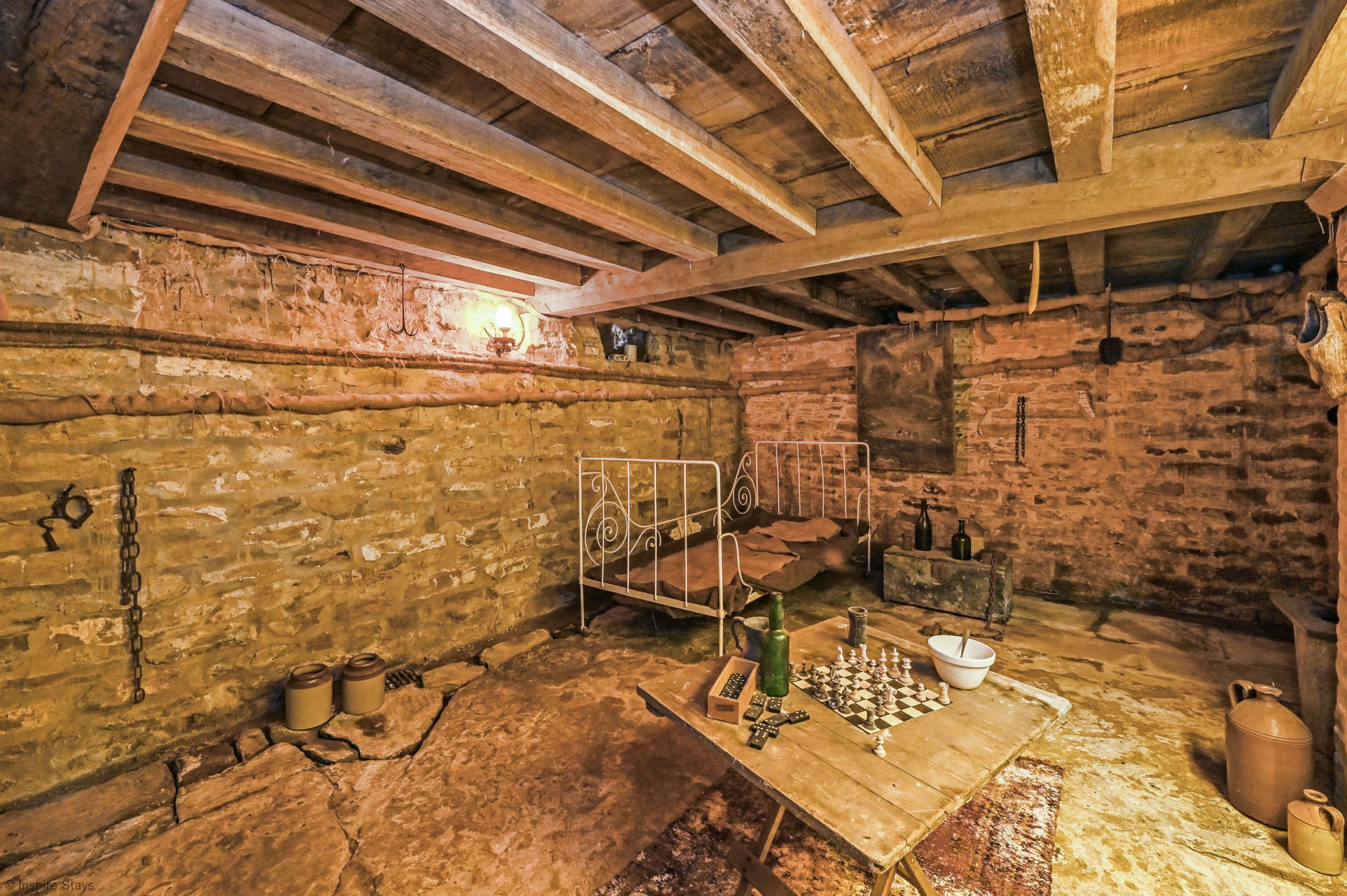 A rustic room with exposed wooden beams, a stone wall, a metal bed frame, and a chessboard on a wooden table.