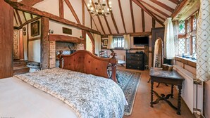 A bedroom with a large bed, wooden beams, a fireplace, a dresser, and a mirror. - Hell Fyre 1594 by inspirestays.com - a cottage that sleeps 2 guests in 1 bedroom (Itchingfield, nr Horsham)