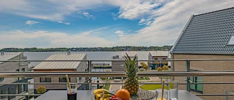 A balcony with a table set for breakfast, including fruits and drinks, overlooking a residential area with modern buildings.