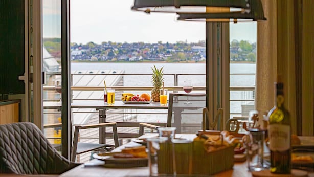 A dining area with a table set for two, a view of a river, and a cityscape in the background. - Wiking-Koje 3 - Elevator, 1st Floor, Underfloor Heating, Shower, Washing Machine (Schleswig)