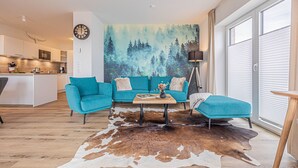 A modern living room with a teal sofa set, a coffee table, and a cowhide rug. A forest mural is on the wall, and there is a kitchen in the background. - Wiking-Koje 3 - Elevator, 1st Floor, Underfloor Heating, Shower, Washing Machine (Schleswig)