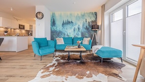 A modern living room with a teal sofa set, a coffee table, and a cowhide rug. A forest mural is on the wall, and there is a kitchen in the background. - Wiking-Koje 8 - Accessible, Elevator, 2nd Floor, Underfloor Heating, Balcony (Schleswig)