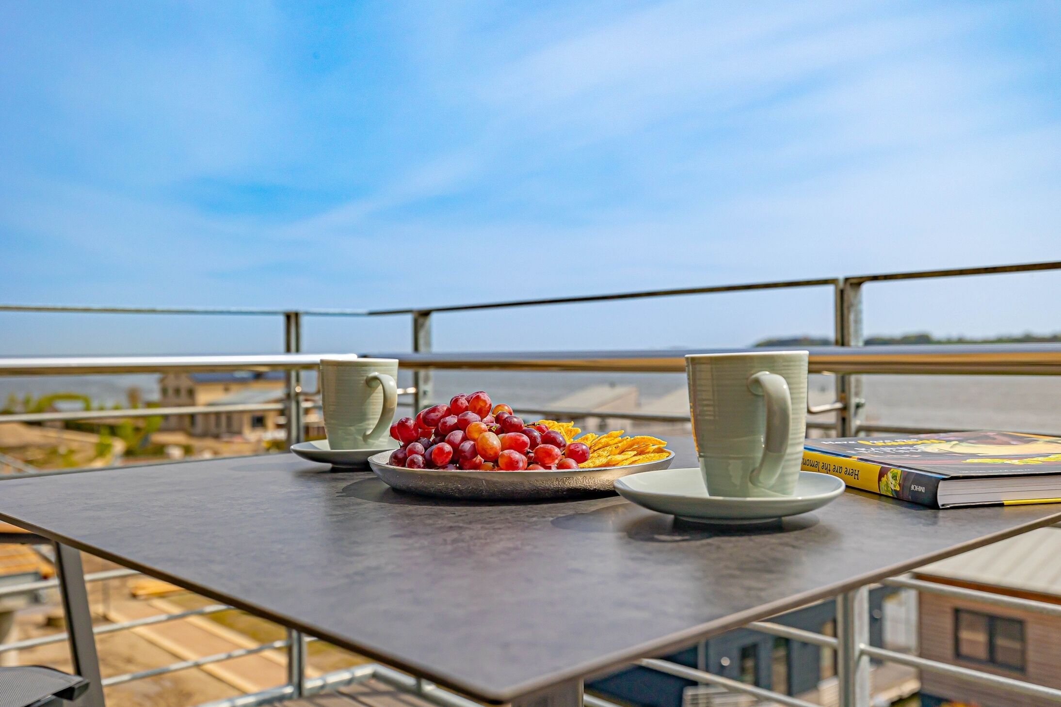 Un balcone con un tavolo apparecchiato per un pasto, inclusa una porzione di frutta e due tazze, con vista sulla città.
