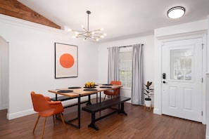 Ein Essbereich mit einem Holztisch, orangefarbenen Stühlen, einer schwarzen Bank, einer Obstschale, einer modernen Kronleuchter und einer weißen Tür.