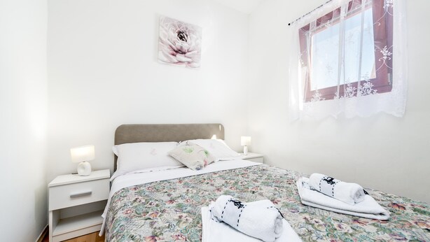 A neatly made bed with floral bedding, a bedside lamp, and a window with a floral curtain. - Studio Nena (Rovinj)