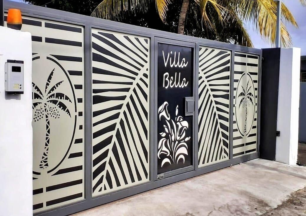 Une entrée fermée par un portail orné d’un panneau métallique décoratif représentant des palmiers et des motifs floraux, et sur lequel est inscrit en lettres noires le nom « Villa Bella ».