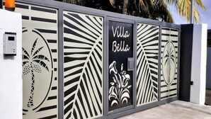 Portilla on koristeellinen metallilevy, jossa on palmu- ja kukkakuvioita, ja mustassa kyltissä lukee nimellä »Villa Bella».