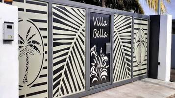 Une entrée fermée par un portail orné d’un panneau métallique décoratif représentant des palmiers et des motifs floraux, et sur lequel est inscrit en lettres noires le nom « Villa Bella ».