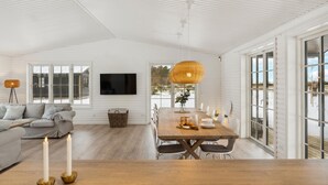 A spacious living room with a large sofa, a dining table with chairs, a TV mounted on the wall, and large windows offering a view of the outdoors.