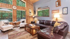 A cozy living room with a leather sofa, a white armchair, and a coffee table. Large windows offer a view of the outdoors.