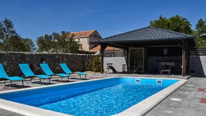 Ein Swimmingpool mit blauem Wasser, umgeben von Liegestühlen und einem überdachten Bereich mit Fitnessgeräten.