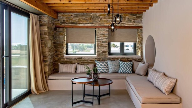 A cozy room with a stone wall, a wooden ceiling, a large window, a sofa, and a round table.