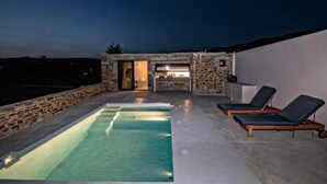 A rooftop pool with lounge chairs, a stone wall, and a small building with an open door.