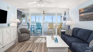 Uma sala de estar moderna com sofá, mesa de centro e televisão. Há uma varanda com cadeiras e vista para o mar.