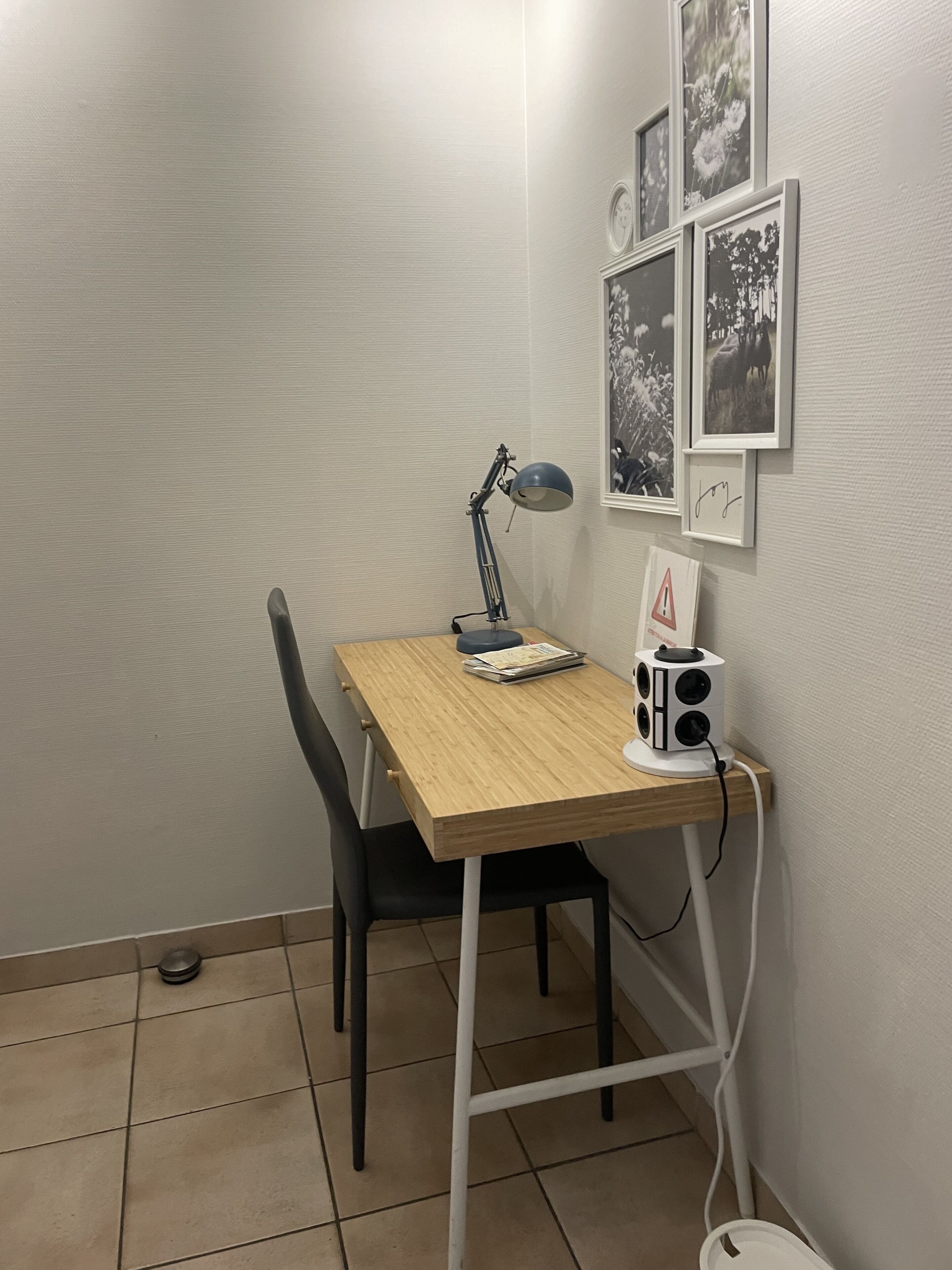 A small study room with a wooden desk, a black chair, a desk lamp, and a white speaker. There are framed pictures on the wall.