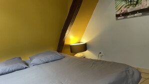 Een bed met grijze beddengoed, een lampje op het nachtkastje en een wand met een plant.